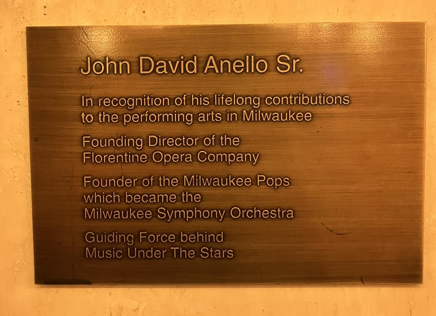 Plaque beneath bust reading - John David Anello Sr. In recognition of his lifelong contributions to the performing arts in Milwaukee. Founding Director of the Florentine Opera Company. Founder of the Milwaukee Pops which became the Milwaukee Symphony Orchestra. Guiding Force behind Music Under the Stars.